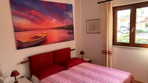 Un dormitorio con cama, almohadas rojas, una mesita de noche, una lámpara, un cuadro de un atardecer y una ventana con cortinas.