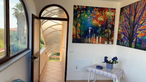 Un pasillo con una puerta de cristal curva que da a un jardín, una pequeña mesa con mantel a cuadros y dos cuadros en la pared.