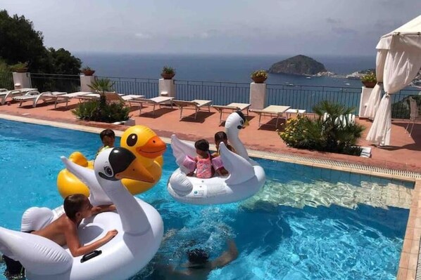 Area piscina con gonfiabili a forma di cigno e anatra, poltrone da relax e vista sul mare.
