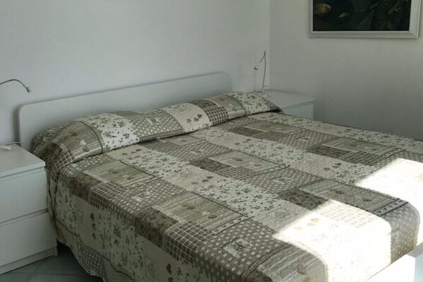 A bedroom with a bed covered in a patterned quilt, a white nightstand, and a wall-mounted lamp. - The "Villa Nunzia" Fig Tree (Barano D'ischia)