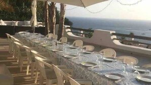 Zona pranzo all'aperto con un lungo tavolo apparecchiato per un pasto, con vista sull'oceano.
