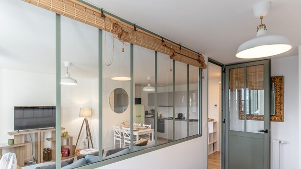 A modern apartment with a large glass wall, a dining area, and a kitchen visible through the glass.