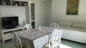 A dining area with a table covered by a floral tablecloth, surrounded by white chairs. A television is mounted on a white cabinet above the table. A white sofa is positioned against the wall, and a painting is displayed above it. - Olive Tree "Villa Nunzia" (Barano D'ischia)