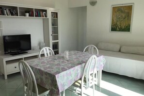 Zona pranzo con tavolo ricoperto da tovaglia a fiori, circondato da sedie bianche. Un televisore è montato su un mobile bianco sopra il tavolo. Un divano bianco è appoggiato al muro, e un quadro è esposto sopra di esso.