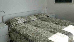 Una camera con un letto singolo, una coperta trapuntata, un comodino bianco e una lampada fissata al muro.