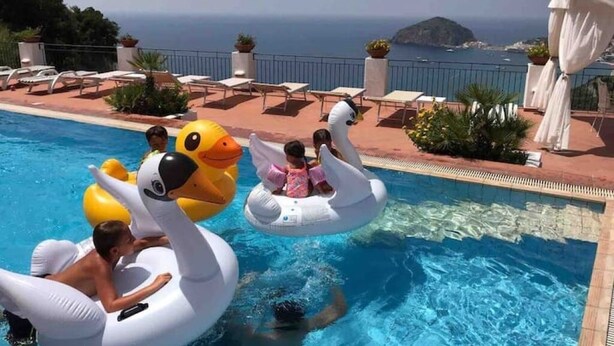 Area piscina con gonfiabili a forma di cigno e anatra, vista libera sul mare e lettini lungo il bordo piscina.