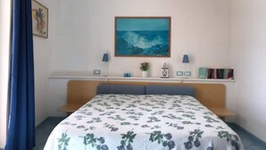 Un dormitorio con cama, mesita de noche, estante con libros, pintura del océano y ventana con cortinas azules.