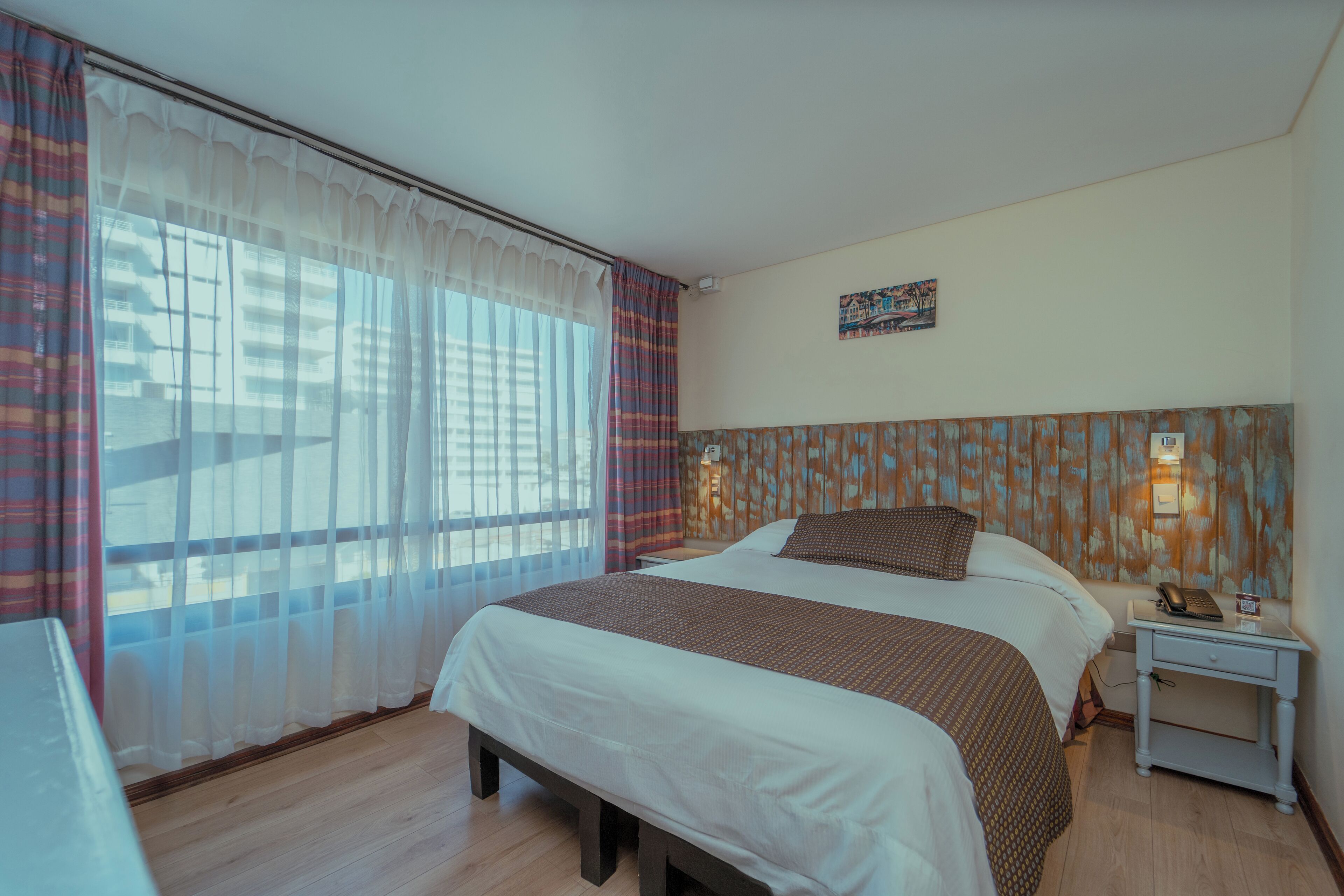 A hotel room with a large bed, a bedside table with a lamp, and a view of the city through sheer curtains.