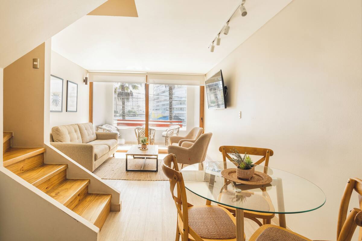 Ein modernes Wohnzimmer mit einem Glastisch, Holzstühlen, einer Couch und einer Treppe, die in eine obere Etage führt.