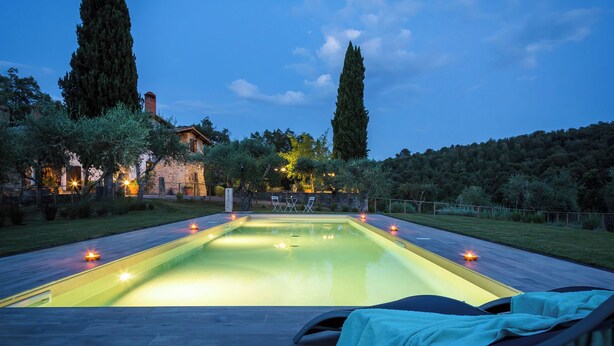 A rectangular swimming pool with a tiled deck, surrounded by trees and a stone building in the background. - Villa Le Liscaie [Crete Senesi]. (Asciano)