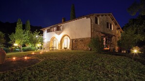A stone building with arched windows and a balcony, surrounded by a well-lit garden at night. - Villa Le Liscaie [Crete Senesi]. (Asciano)