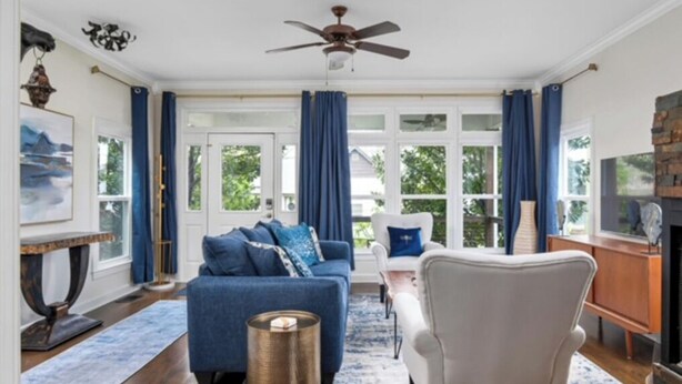 Een ruime woonkamer met een blauwe bank, een witte fauteuil en een salontafel. Er is een plafondventilator, een open haard en een groot raam met blauwe gordijnen.
