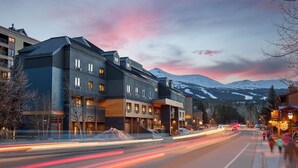 Un hotel de varios pisos con diseño moderno, que presenta una combinación de fachadas de madera y metal, situado en una zona montañosa con vistas a picos nevados.