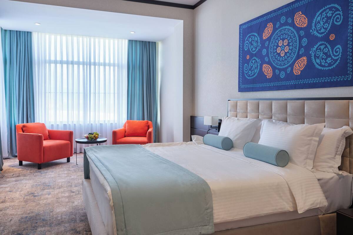 A hotel room with a large bed, two armchairs, a small table with a fruit bowl, and a large abstract painting on the wall.