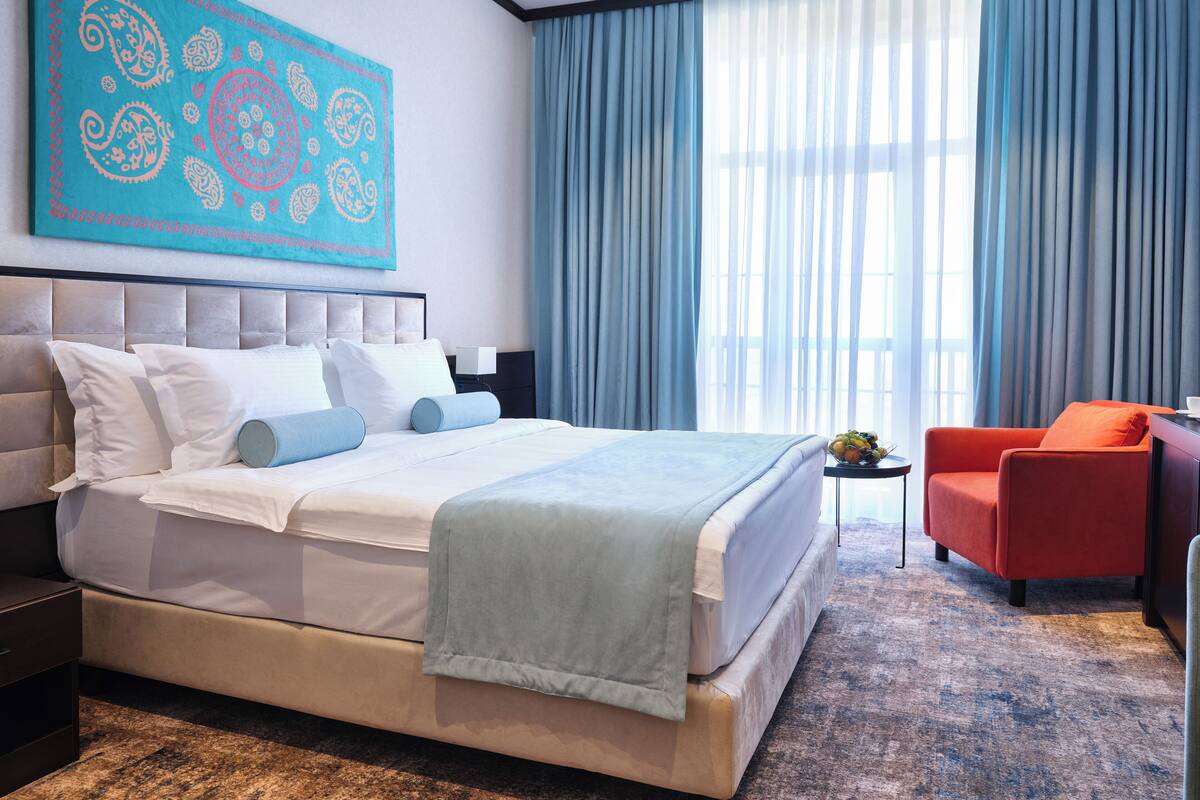 A modern hotel room with a large bed, a red armchair, and a large window with blue curtains.