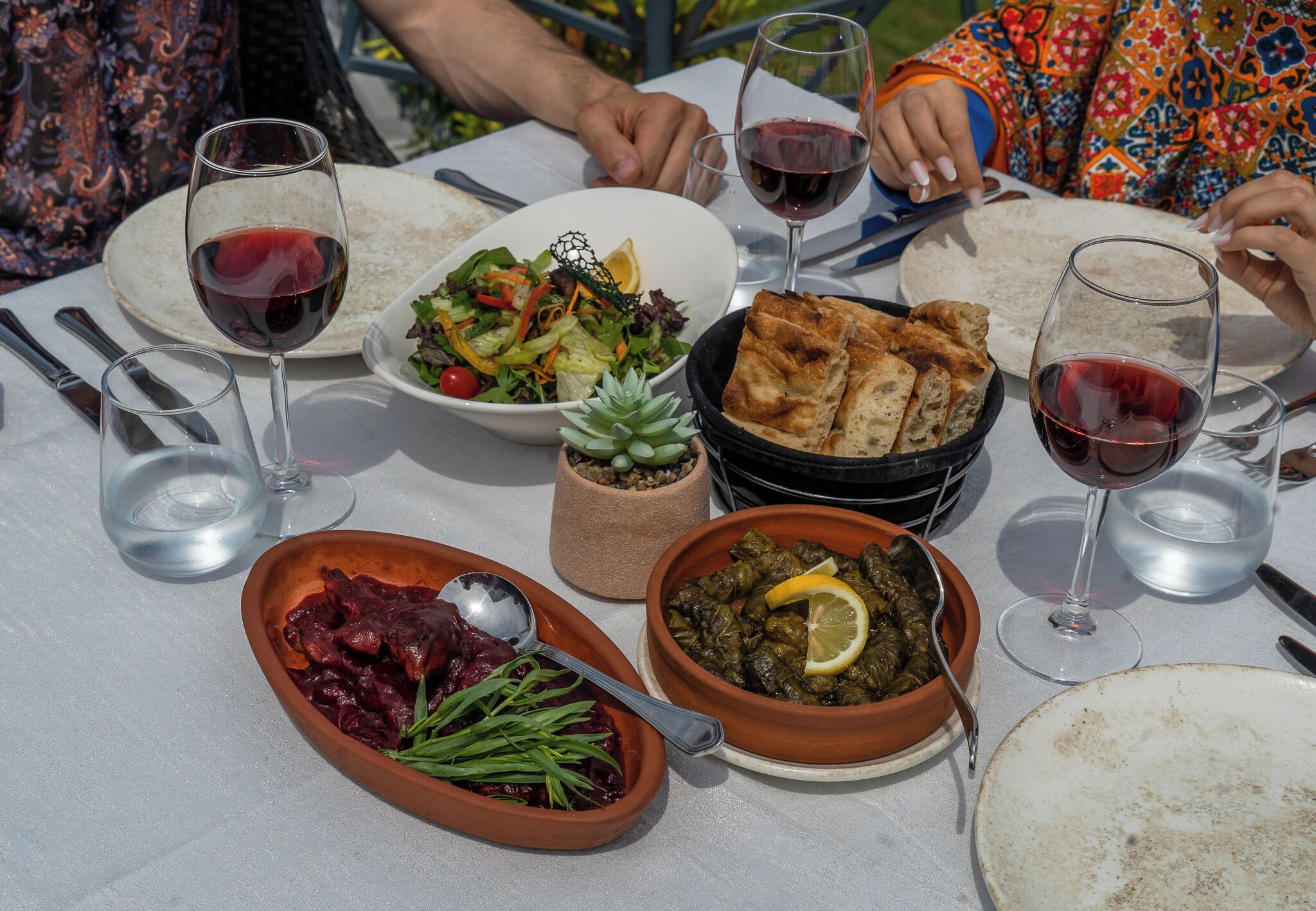 Una mesa dispuesta con diversos platos, como una ensalada, hojas de parra rellenas y un bol de pimientos rojos, junto con copas de vino tinto.