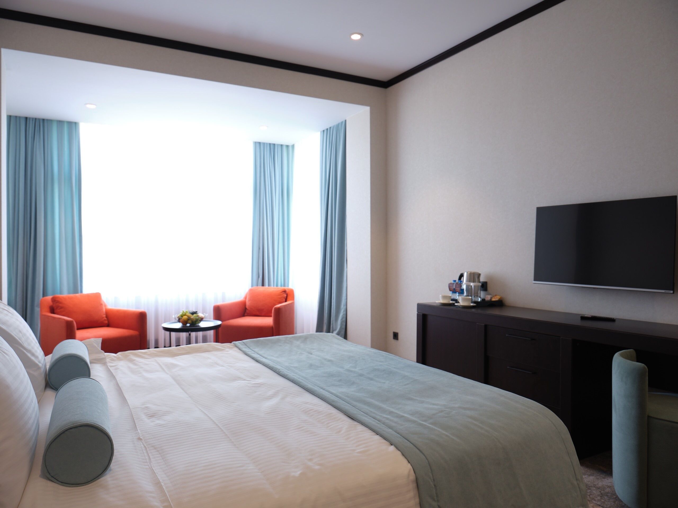 Habitación de hotel con una cama grande, dos sillones, una mesita con frutero, cafetera y televisor de pantalla plana en la pared.