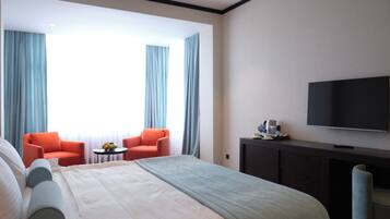 Habitación de hotel con una cama grande, dos sillones, una mesita con frutero, cafetera y televisor de pantalla plana en la pared.