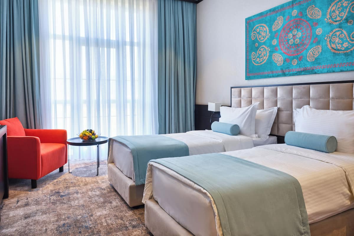A hotel room with two beds, a red armchair, a small table with fruit, and a large abstract painting on the wall.