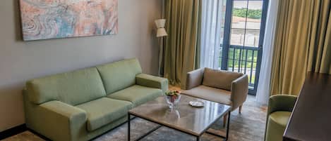 A living room with a green sofa, a beige armchair, a glass coffee table, and a painting on the wall.