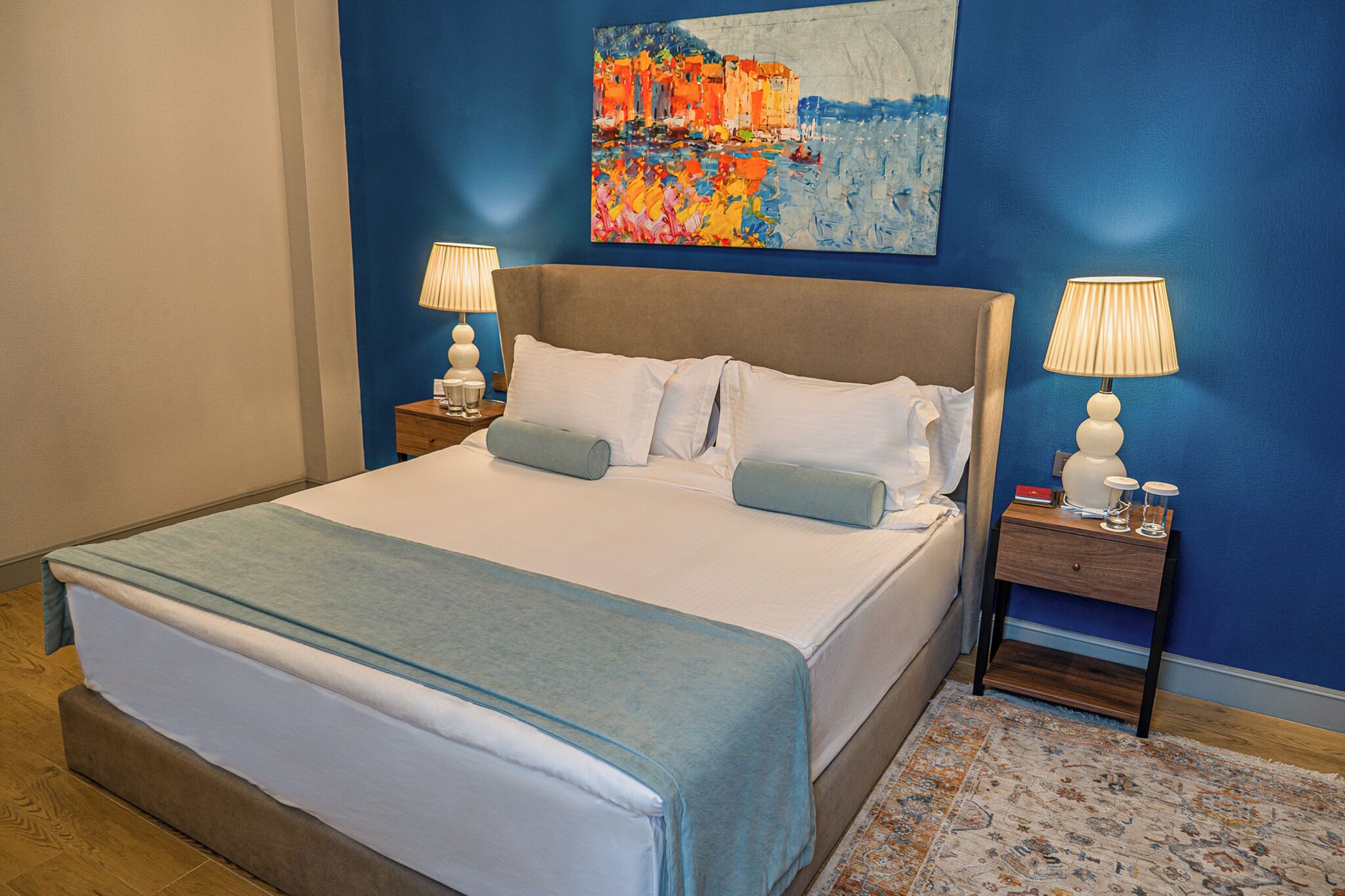A neatly made bed with a blue blanket, two bedside lamps, and a painting above the bed.