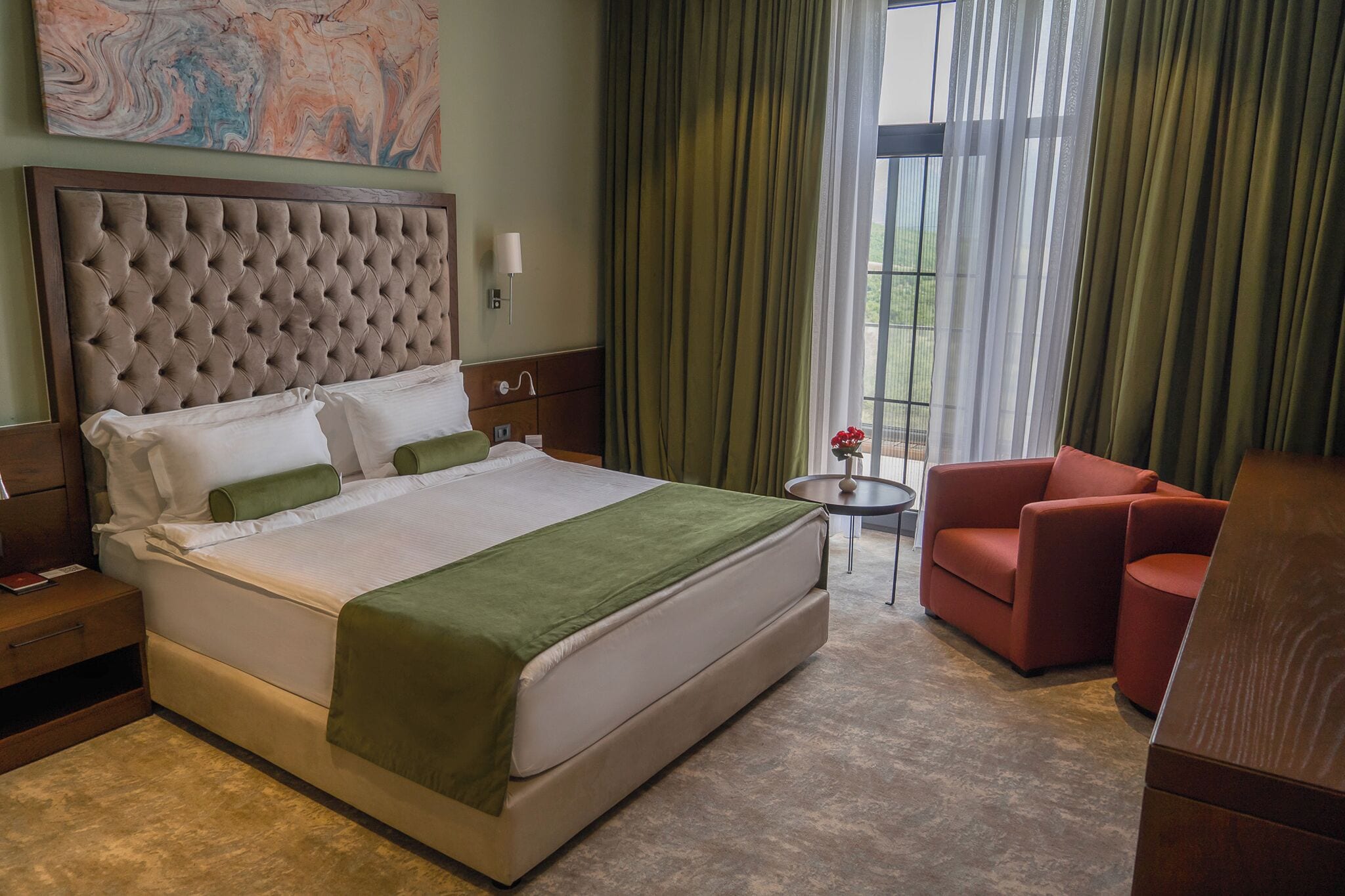 Una habitación de hotel con una cama grande, una mesita de noche, un sillón rojo, una pequeña mesa y una ventana con cortinas.