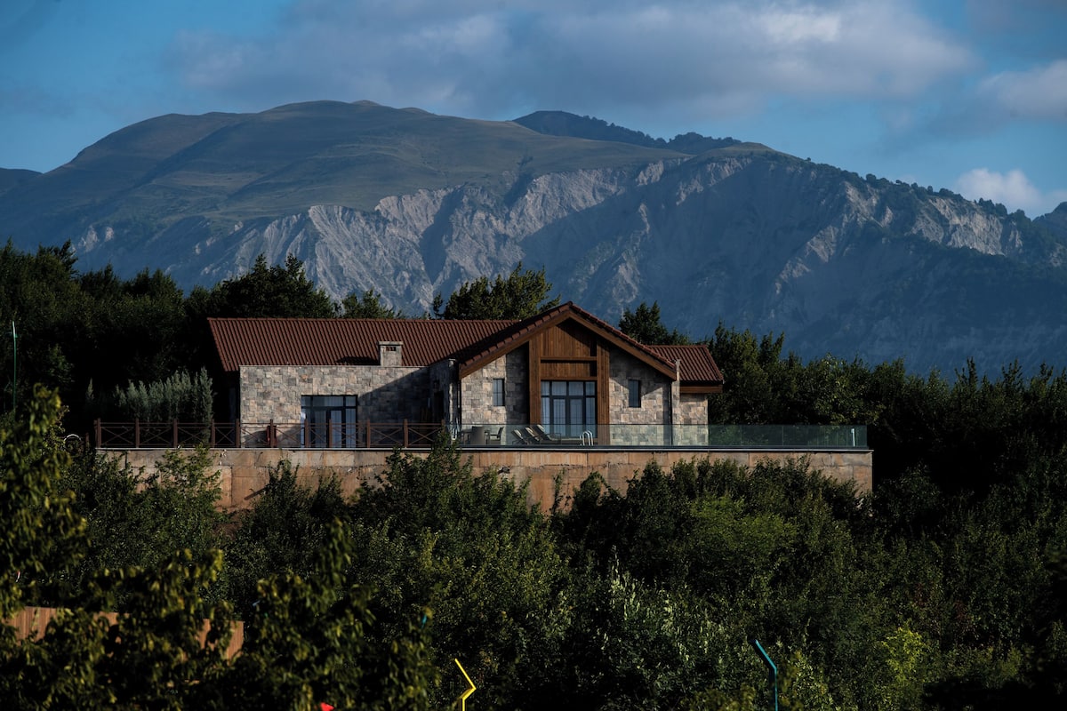A stone house with a wooden roof, set against a mountainous backdrop.