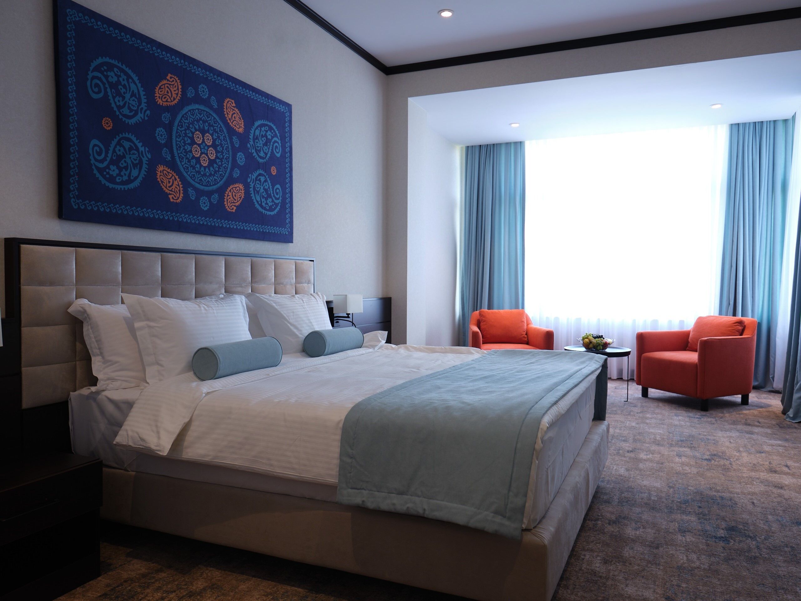 A modern hotel room with a large bed, two chairs, a desk, and a large window with curtains.