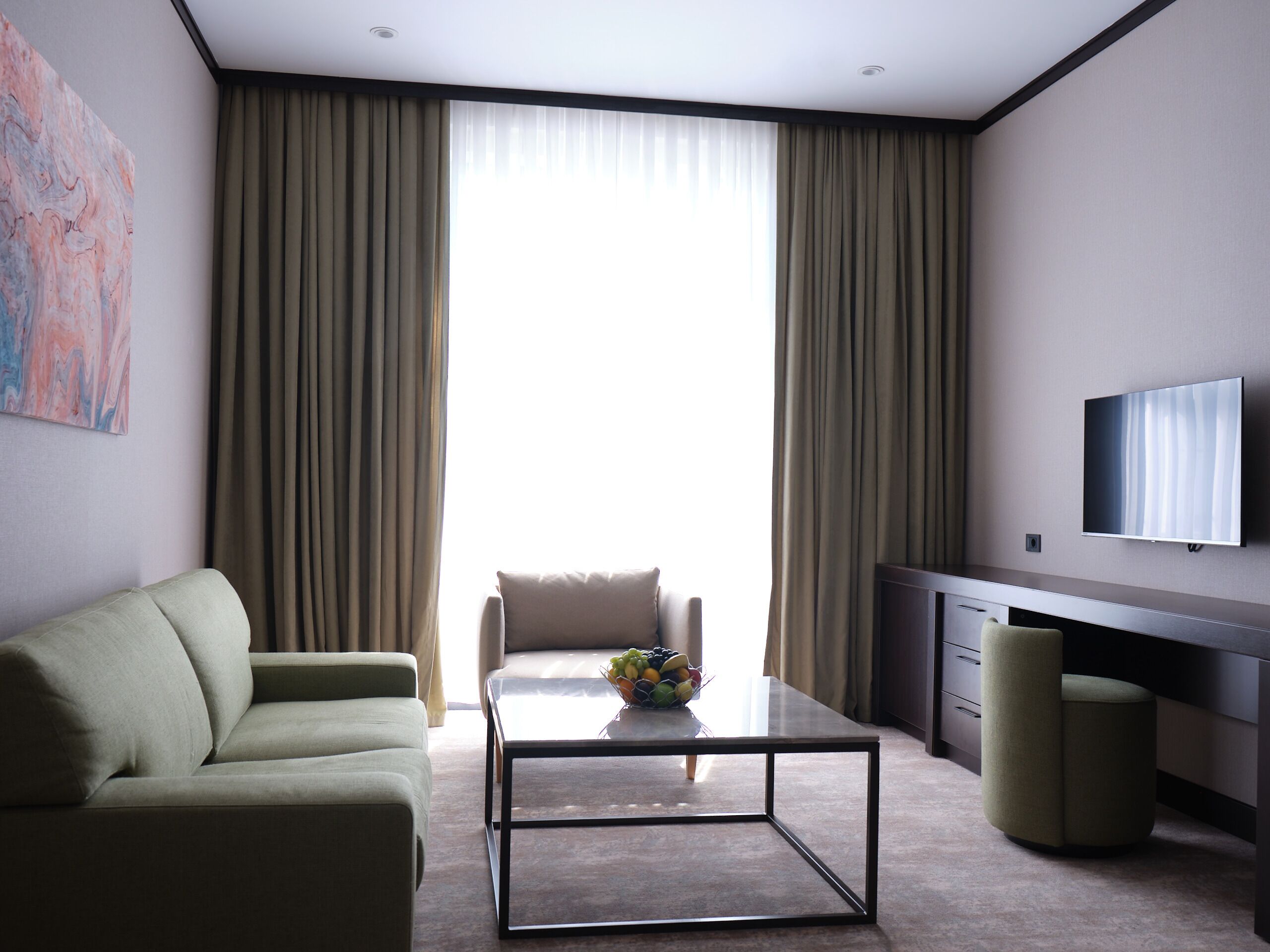 A modern hotel room with a sofa, a coffee table, a TV, and a large window with curtains.