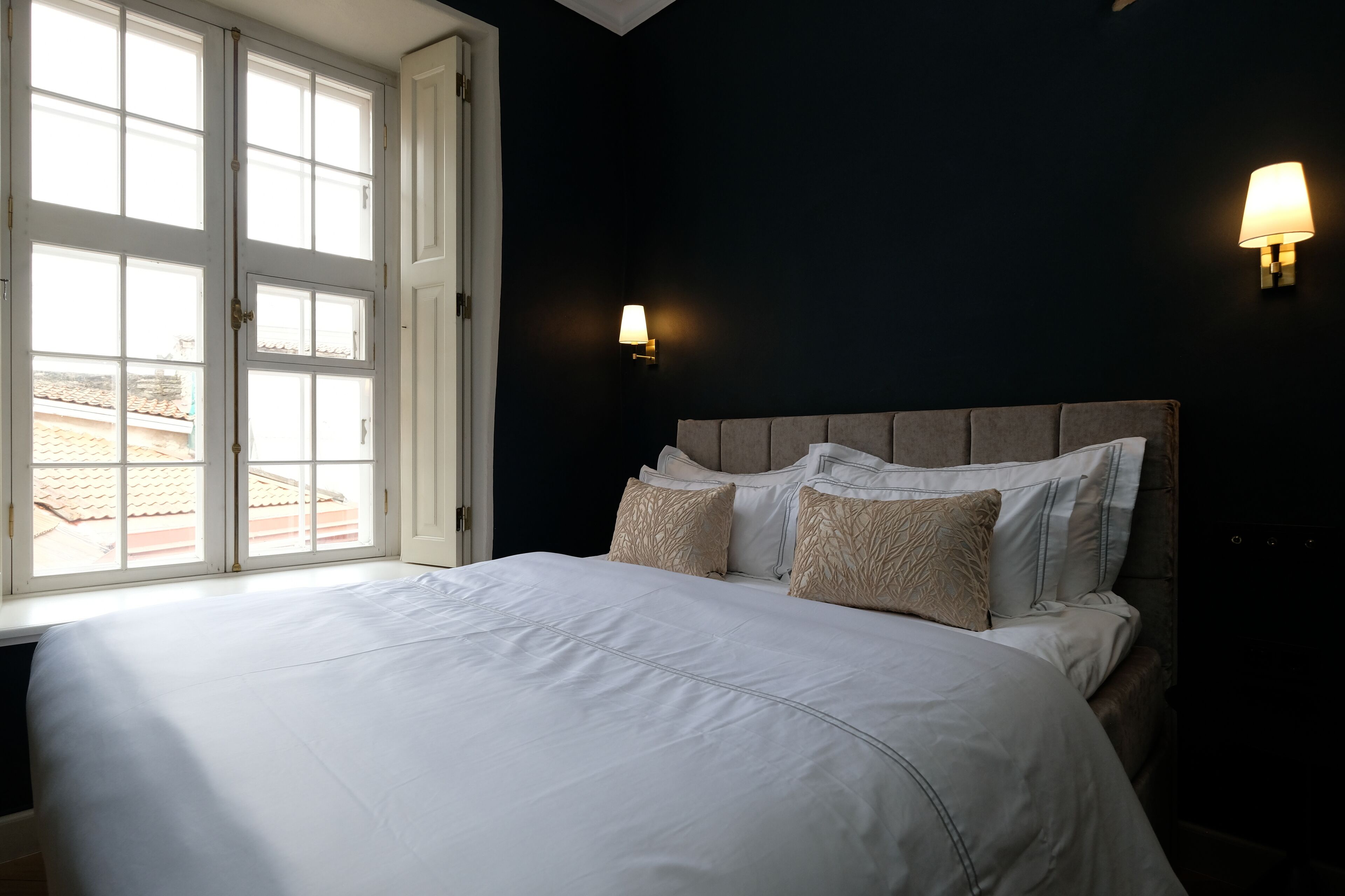 A bedroom with a large bed, two wall-mounted lamps, and a window with a view of buildings.