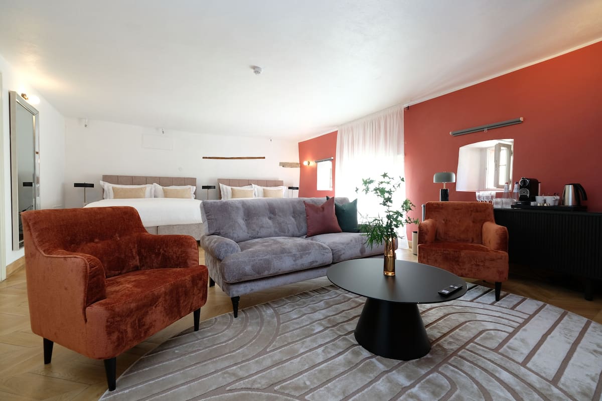 Moderni hotellihuone, jossa on harmaa sohva, kaksi punaista nojatuolia, musta sohvapöytä ja valkoisilla lakanoiden päällä varustettu sänky.
