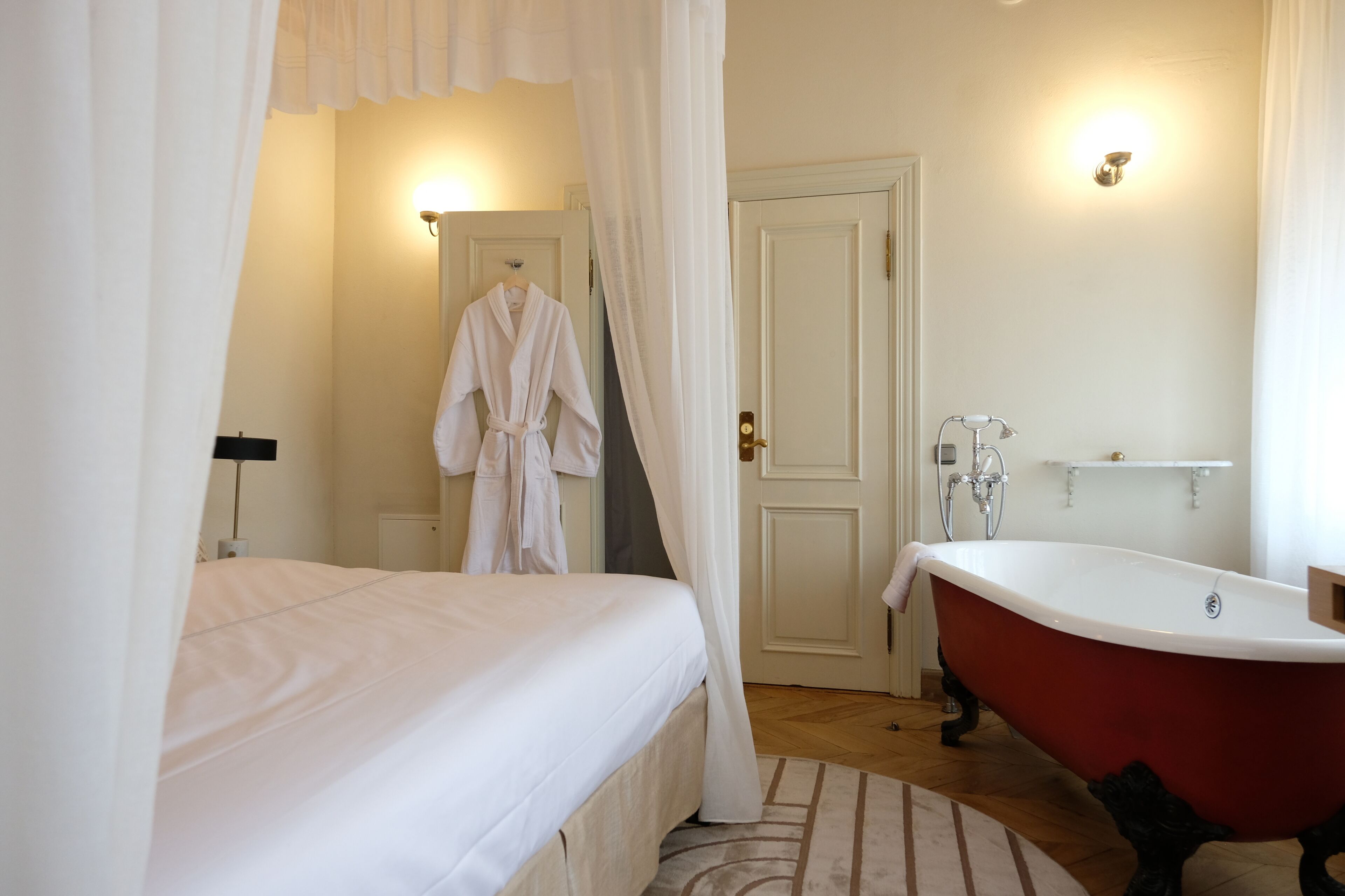 A bedroom with a bed, a red clawfoot bathtub, and a white robe hanging on a door.