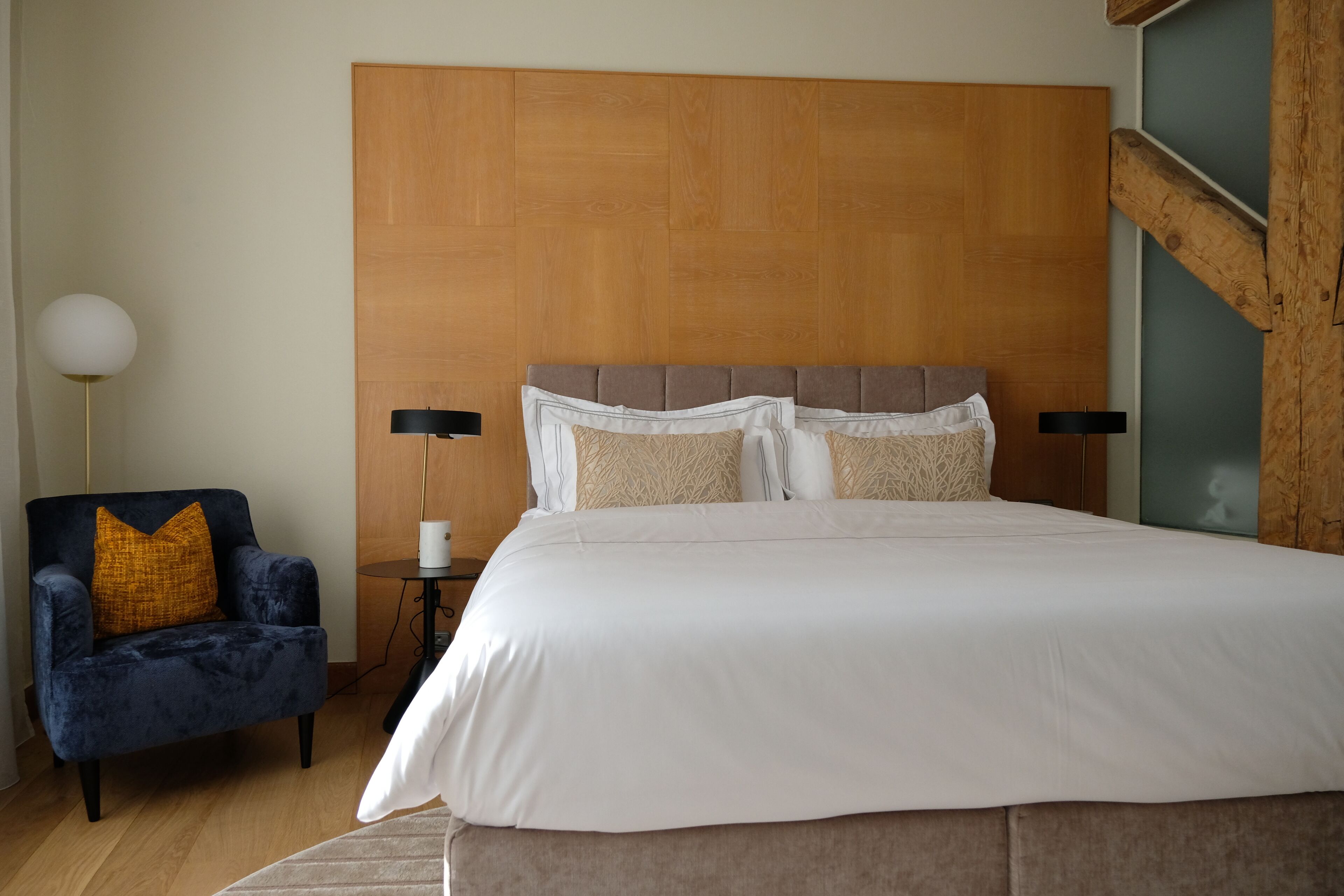 A bedroom with a large bed, a dark armchair with a yellow pillow, a bedside table with a lamp, and a wooden headboard.