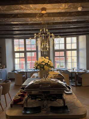 A dining area with a buffet setup, a chandelier, and large windows with a view of buildings. - The Three Sisters Hotel (Tallinn)
