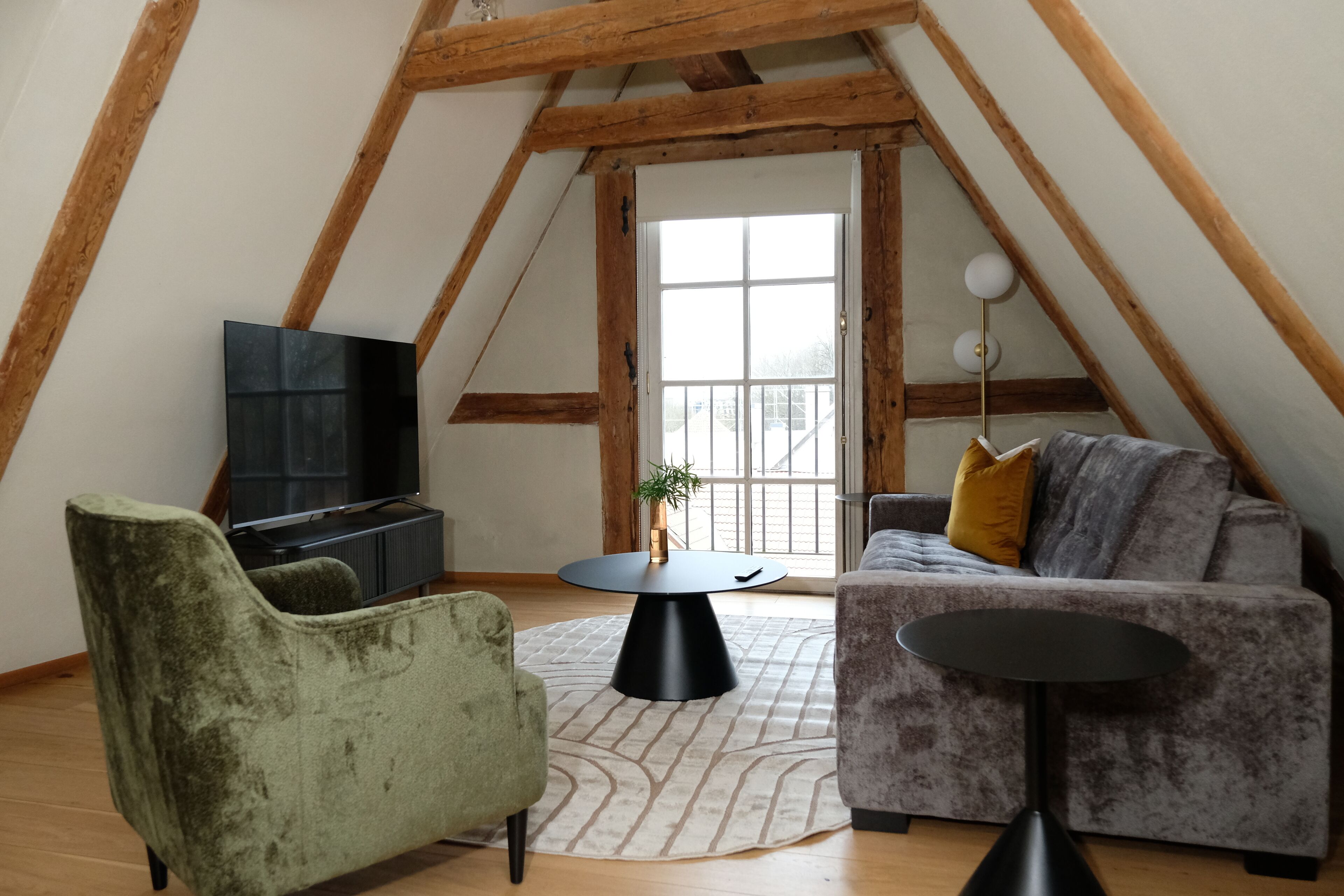 An attic room with a wooden ceiling, a television, a green armchair, a grey sofa, and a small table.