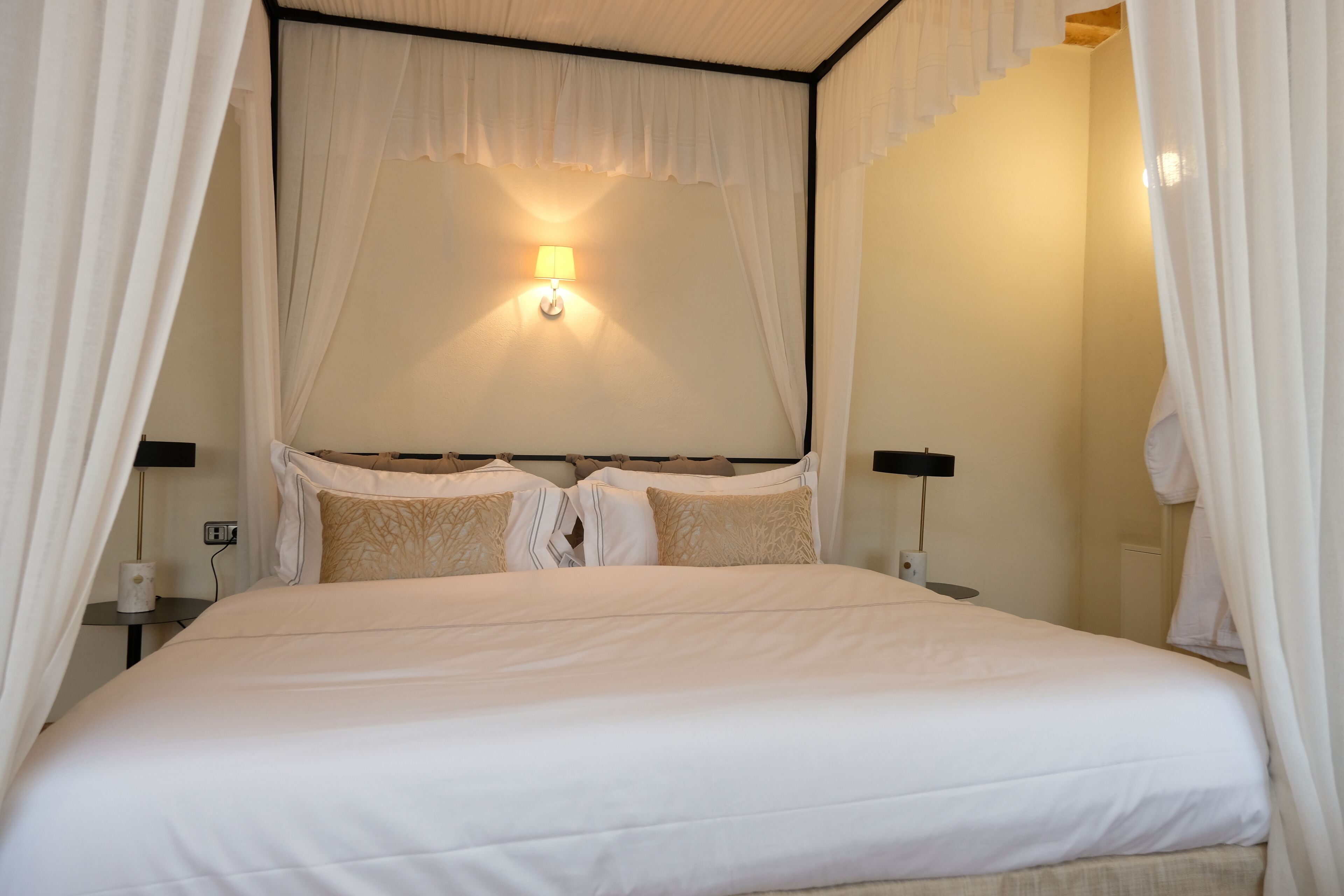 A neatly made bed with white bedding and a canopy, a bedside lamp, and a wall-mounted light fixture.