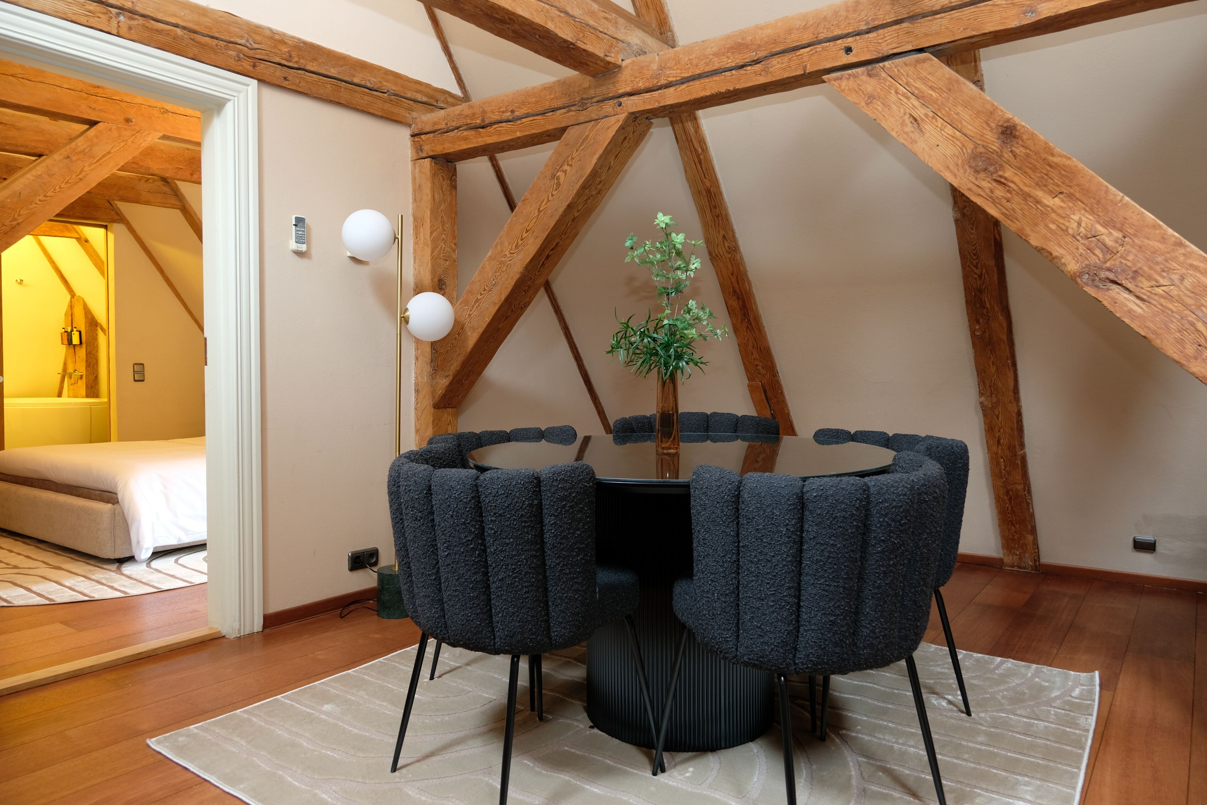 Un comedor con una mesa redonda y dos sillas, vigas de madera en el techo, una planta en maceta y una cama al fondo.