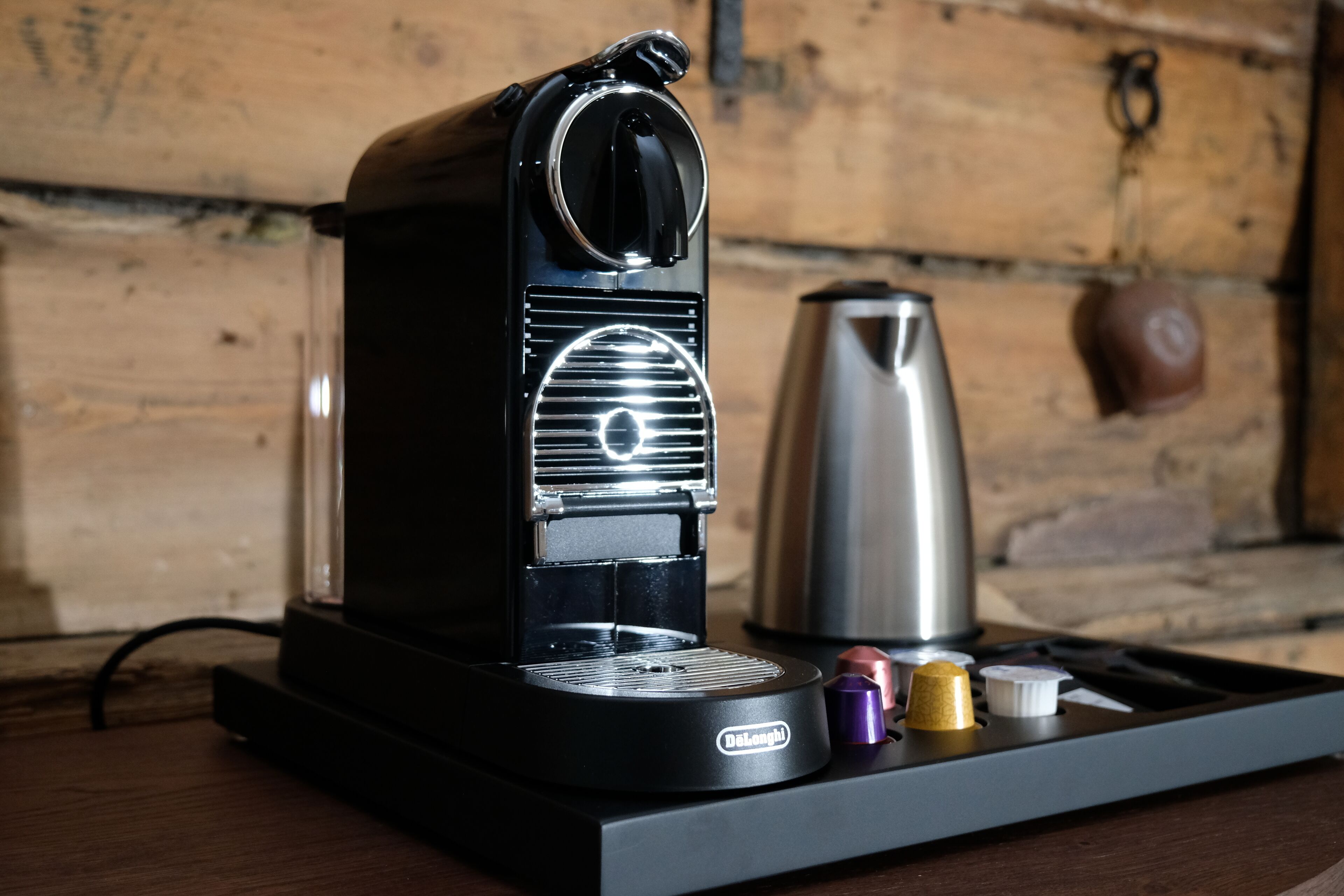Una cafetera DeLonghi sobre una bandeja negra, con una jarra de acero inoxidable y una taza marrón al fondo.