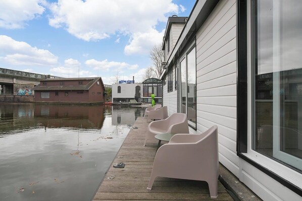 Un edificio a orillas del río con una terraza de madera, sillas y una persona con chaqueta verde.