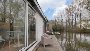 A modern houseboat with large windows, a wooden deck, and outdoor seating by the water. - HouseBoat the NEW LAKE (Amsterdam)
