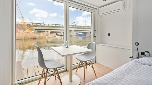 A modern hotel room with a large window, a bed, a table with two chairs, and a view of a bridge over a river. - HouseBoat the NEW LAKE (Amsterdam)