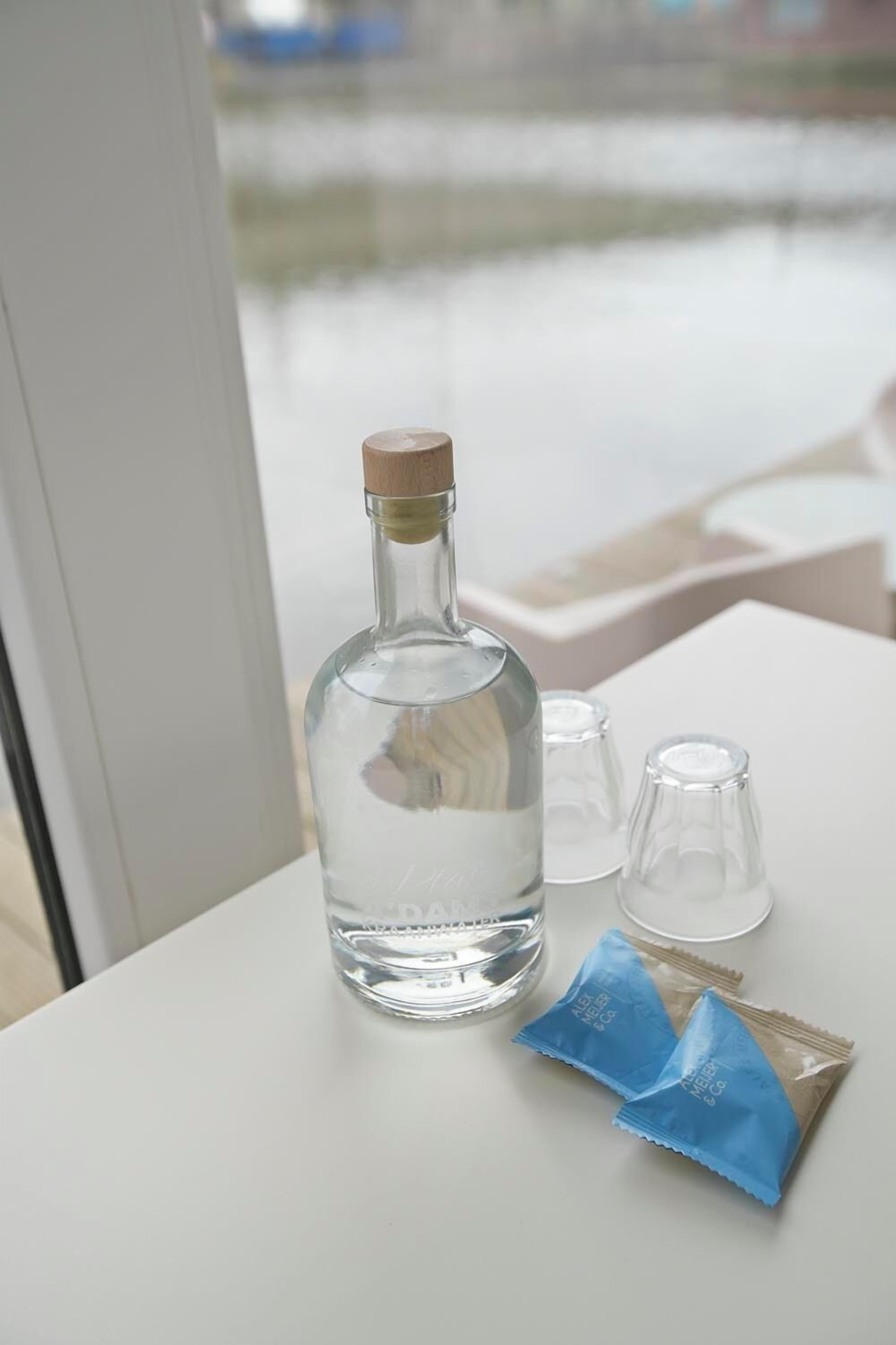 A bottle of water, two glasses, and two packets on a white table by a window.