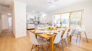 Zona de menjador moderna amb taula de fusta preparada per a dinar, cadires blanques i vista a la cuina al fons.