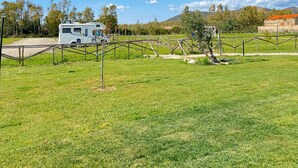 A grassy field with a white RV parked near a fence, a stone building in the background, and trees under a clear sky. - Munai Sardinia (San Giovanni Suergiu)