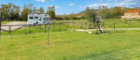 A grassy field with a white RV parked near a fence, a stone building in the background, and trees under a clear sky.