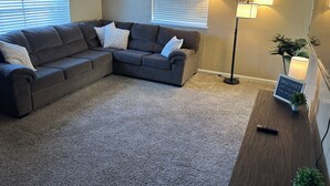 A living room with a sectional sofa, two floor lamps, a small table with a sign, and a potted plant.