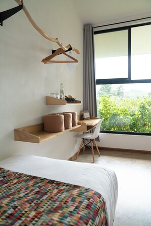 A hotel room with a bed, a desk, a chair, a window with a view of greenery, and a wooden shelf with items. - Hotel con Corazón Monteverde (Monteverde)