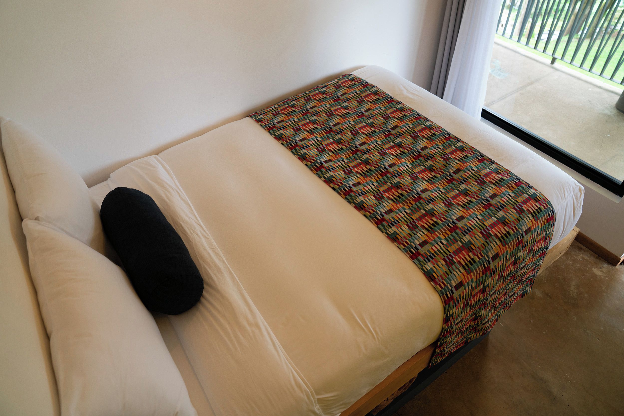 Ein Einzelbett mit einer gemusterten Decke und einem schwarzen Kopfkissen in einem Zimmer mit Blick ins Freie.