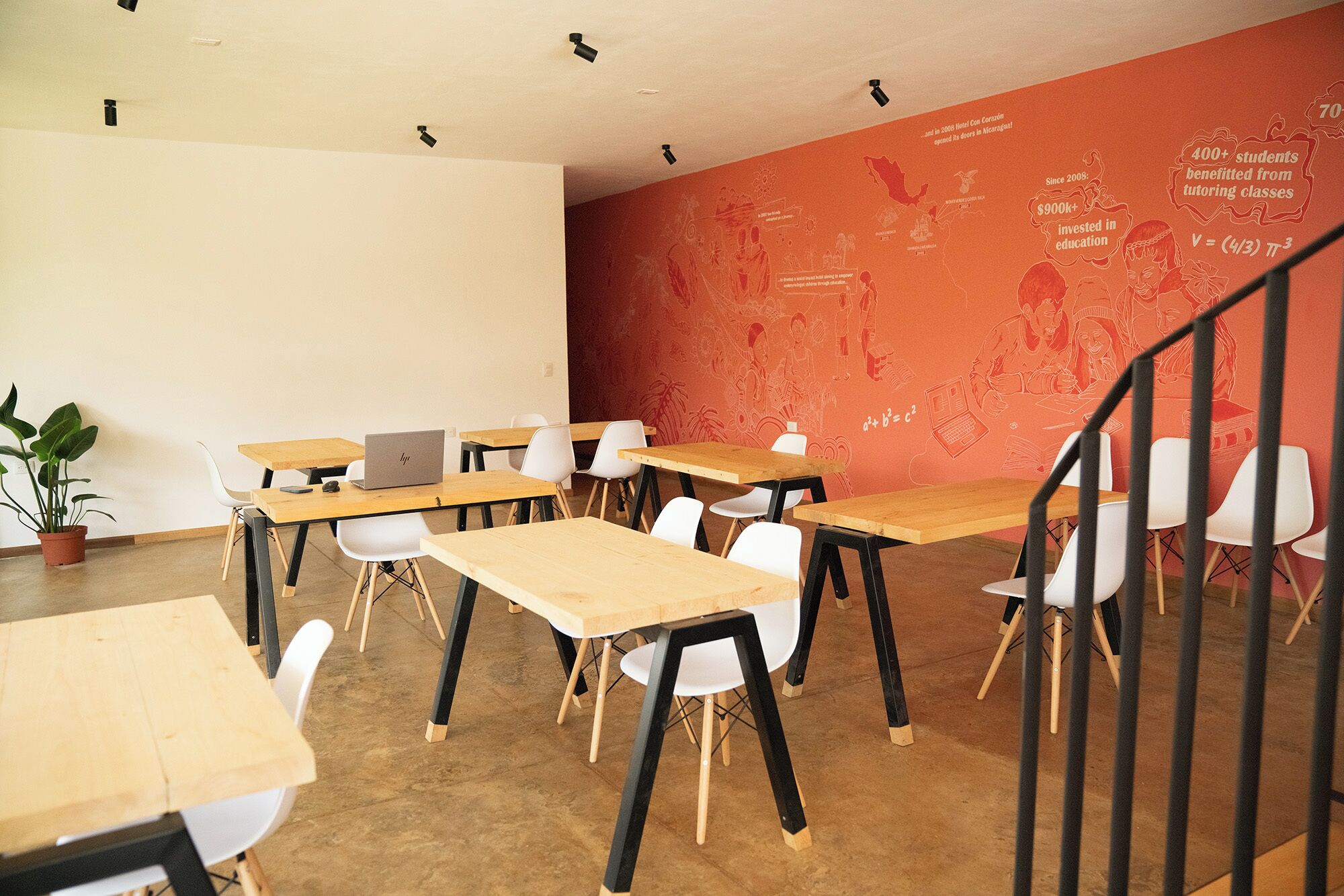A classroom with wooden tables and white chairs, a laptop on one table, and a red wall with educational graphics.
