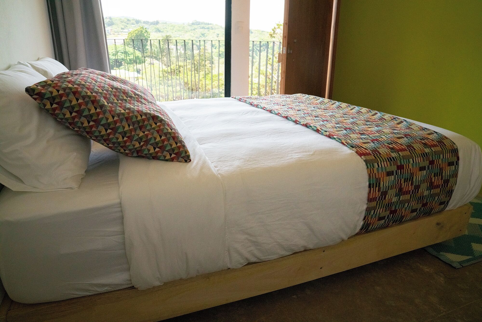 A bed with white linens and a colorful geometric-patterned pillow, set against a green wall with a view of greenery outside.